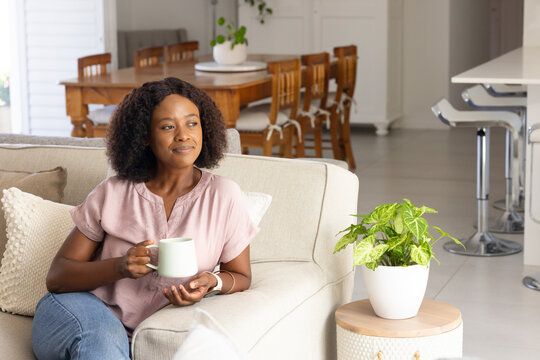 African woman sitting on light beige sofa holding white mug in living room with throw pillows