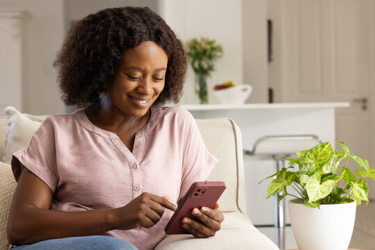 Adult woman sitting on sofa at home, tapping smartphone after notification, replying to friend