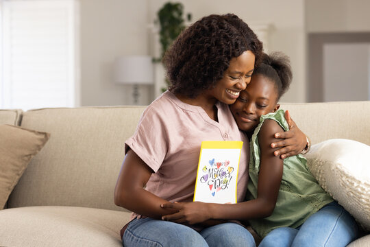 African American mother and child holding Mother's Day card, reading message and hugging on sofa