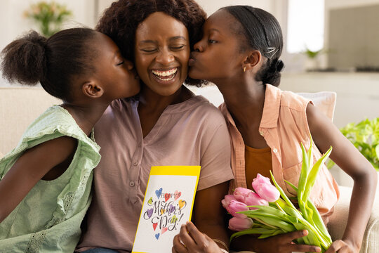 African American mom with youth and child girls laughing on sofa holding Mother's Day card, tulips