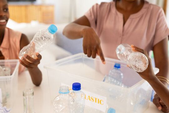 Pointing adult wearing pale pink top guiding two girls sorting bottles at home, with plastic bin