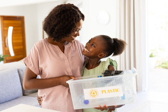 African American woman and girl holding bin labeled PLASTIC at home, sorting plastics for recycling