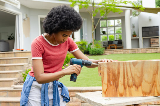 Blue cordless drill is driving screw into plywood box on worktable in backyard, with stone steps