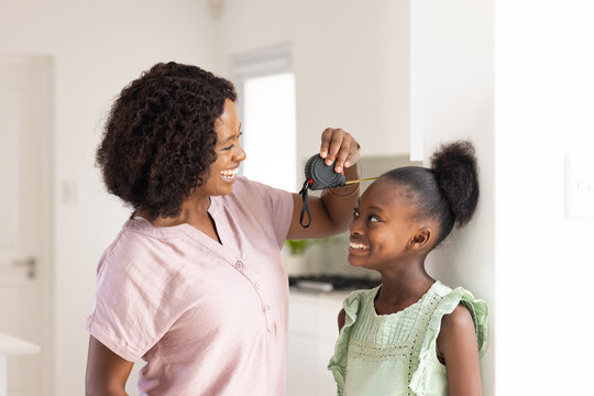 Measuring mother in pink top holding tape measure above girl in green dress at home kitchen