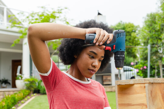 African American woman wearing red tee dark headband using drill on wooden crate in backyard