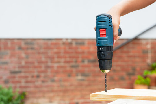 DIYer holding cordless drill raising over wooden plank on patio bench near brick wall, copy space