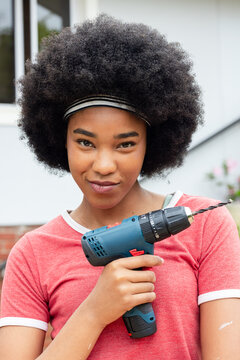 African woman holding blue drill with long bit, wearing red tee, smiling outside house