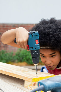 Blue cordless power drill is driving screw into light wooden plank on workbench in backyard