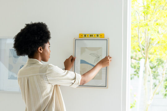 African American woman hanging framed abstract print at home using yellow spirit level by window