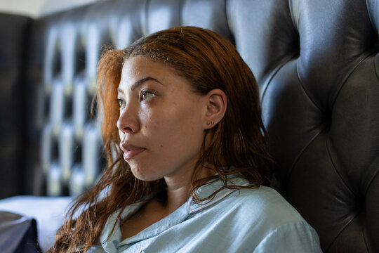 Sitting woman wearing light blue button shirt gazing left on bed, with tufted leather headboard