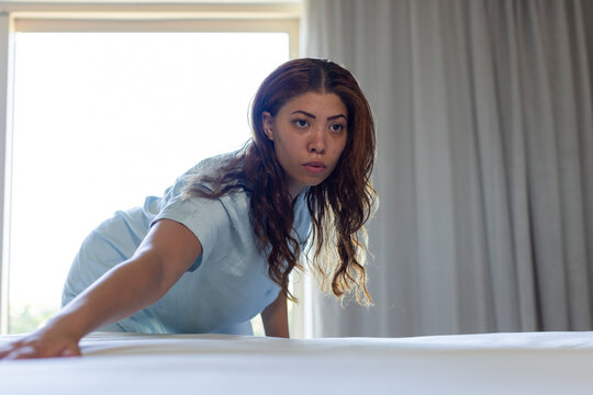 African American woman wearing blue uniform smoothing and tucking white sheet on bed in hotel room