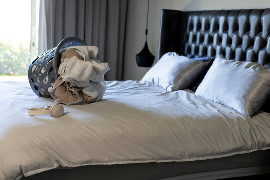 Showing bed with light gray duvet rumpling in bedroom, with tipped laundry basket spilling towels