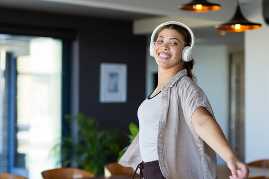 Smiling woman dancing in modern living dining area, wearing white headphones and beige shirt
