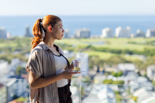 Standing on balcony, woman wearing white tank, beige shirt holding glass cup with lemon, copy space