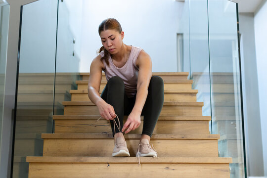 African American woman sitting on stairs tying light shoes near glass railing wearing tank leggings