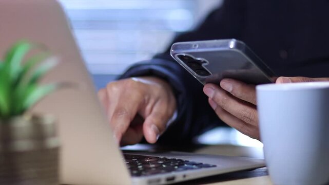 Close up of a man's hands typing a password on a laptop keyboard and using a smartphone for two factor authentication, ensuring data security and online privacy