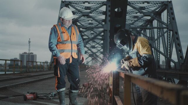Two construction workers in safety gear repair a metal bridge railing. One welds with sparks while the supervisor watches. Ideal for industrial, safety, and infrastructure projects.