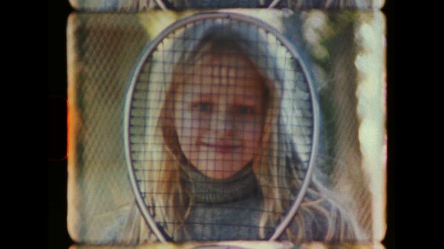 Authentic vintage home movie from the 1970s. A young blonde girl smiles while holding a tennis racket and ball on a sunny court. Nostalgic, grainy Super 8 film look with light leaks.