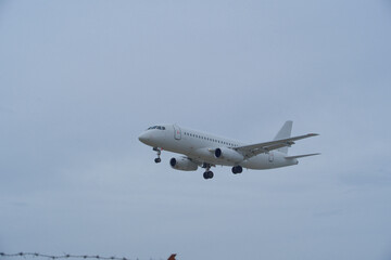 Obraz premium White Plane Approaching Runway Behind Barbed Wire Fence. White Commercial Airplane Approaching Runway Behind Barbed Wire Fence on Overcast Day