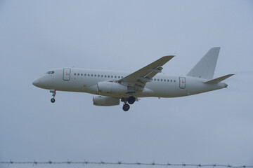Obraz premium White Plane Approaching Runway Behind Barbed Wire Fence. White Commercial Airplane Approaching Runway Behind Barbed Wire Fence on Overcast Day