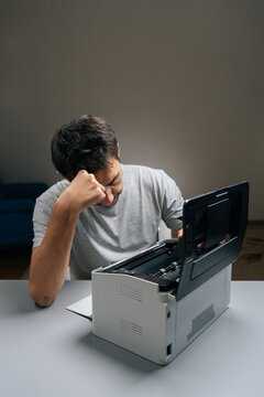 Frustrated man sitting at desk with paper jam in malfunctioning office printer, troubleshooting and trying to fix device amid stress and deadline pressure in home office setting.