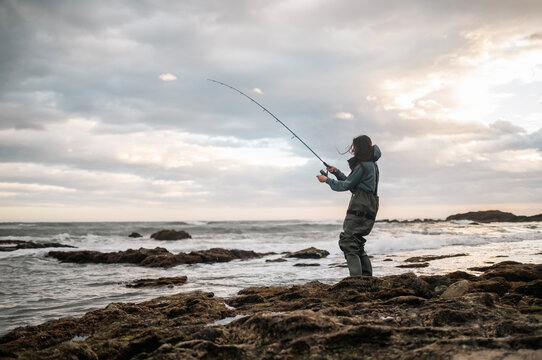 Woman Catching Fish from Rocky Shore with Spinning Rod, Fishing Success, Outdoor Sport, Coastal Adventure, Female fisherwoman on the beach fishing during a stormy day