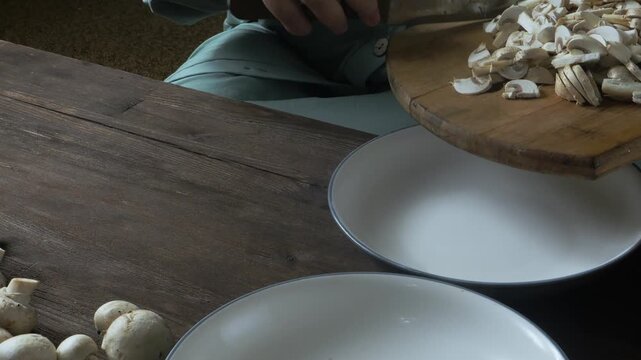 A woman's hands slice champignons with a knife on a cutting board on the table and place them on a plate. Medium shot.