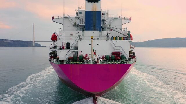 Bulk carrier with vivid magenta hull navigating Bosphorus Strait with suspension bridge in background at dusk. Close up, Aerial tracking shot