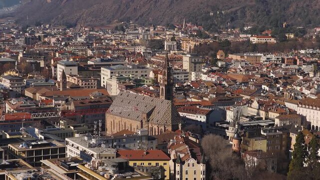 Aerial view of Bolzano, Italy, with a Gothic cathedral, diamond tiled roof, tall spire, terracotta rooftops, a red crane, and Alpine slopes in crisp sunlit tones.