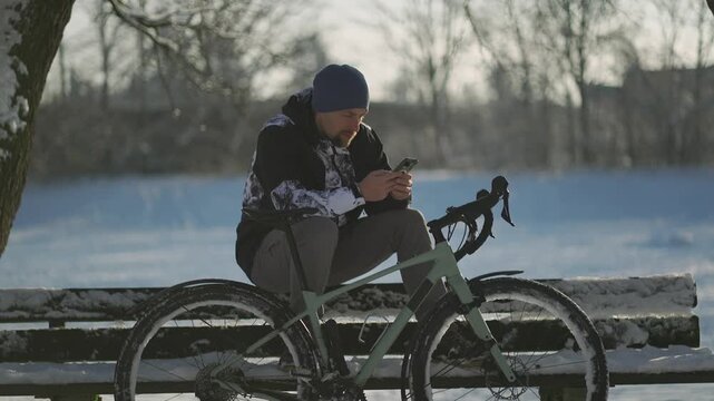 A winter bicycle commuter checks a smartphone map for navigation while resting on a snowy bench. Staying connected and active during a cold bike ride through a German city park in any weather.