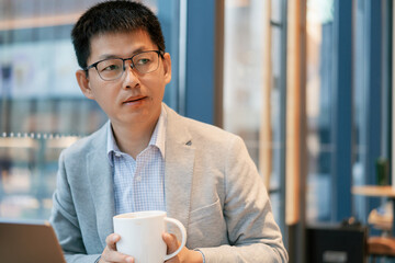 A middle-aged East Asian man wearing glasses holds a white mug in a modern office setting