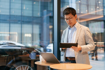 Fototapeta premium Businessman in a light suit holding a tablet while reviewing data near a window in a modern café