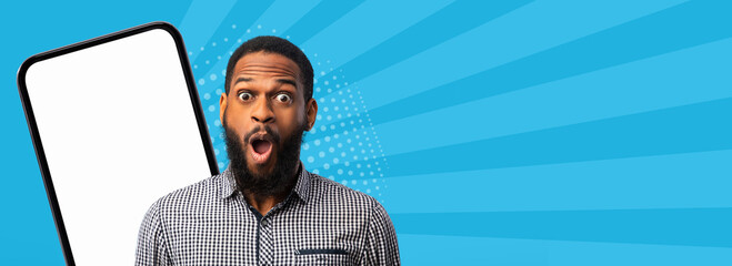 A man stands with a shocked expression, looking at a blank smartphone screen. The background has a bright design with blue rays. This scene captures a moment of surprise. © Prostock-studio