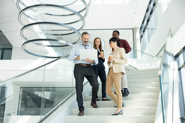 Diverse team of professionals walking down a modern office staircase, collaborating over a laptop and discussing a project while smiling © Dejan