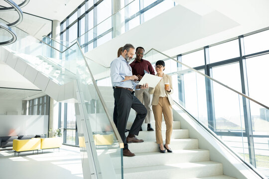 Diverse business team collaborating and discussing work using a laptop while standing on modern office stairs