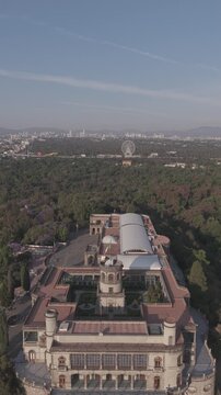 Vertical Aerial view over Chapultepec Castle at sunrise with a panoramic view of Bosque de Chapultepec and the Aztl&aacute;n Ferris wheel visible in the distance under a clear blue sky.