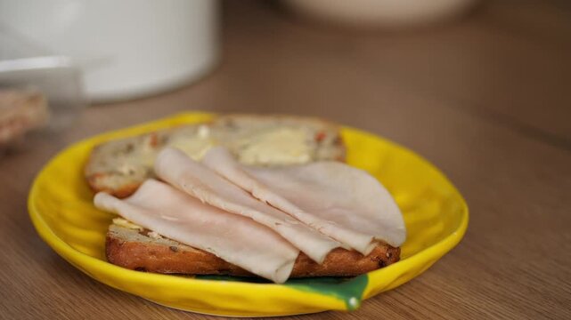Video series sandwich. part 4, Hands placing a slice of sausage onto fresh bread that rests on a bright yellow plate for breakfast or a snack. The image captures a close-up, 