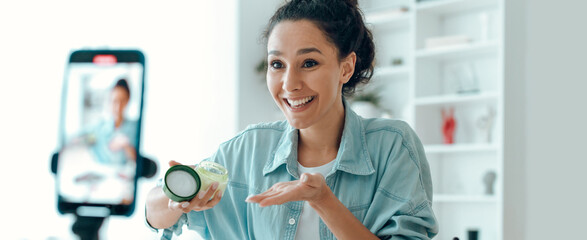 A woman is smiling and holding a skincare jar while talking to a camera in a home environment. She...