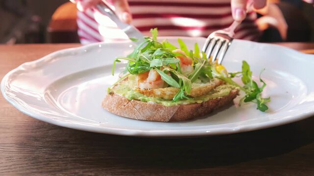 Close-up of woman eating gourmet bruschetta with guacamole, shrimp, arugula and poached egg in restaurant. Stylish brunch appetizer and modern gastronomy concept.