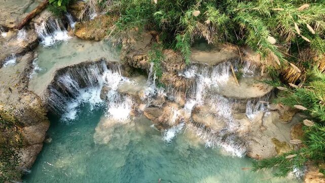 Aerial drone view of turquoise pools and cascading limestone waterfalls at Kuang Si Falls, flowing through rock terraces and jungle vegetation in northern Laos