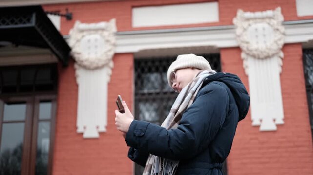 Teenager Girl Walking Alone on City Street with Smartphone. Portrait of 14 years old girl wearing eyeglasses, wool hat, winter jacket using mobile phone. Urban Style. Rainy mood weather. Cold season