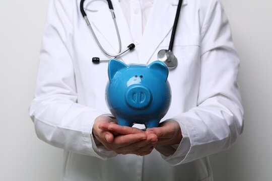 Doctor in medical coat with blue piggy bank and stethoscope on light grey background, closeup