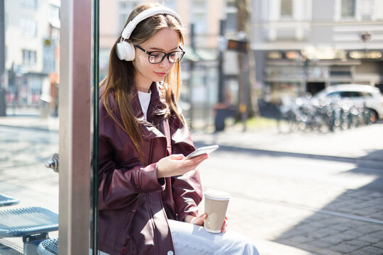 Girl sitting at tram station. City commute, urban lifestyle. Student waiting for public transport. Young woman with smartphone, headphones, coffee scrolling outdoors, doomscrolling, mobile addiction