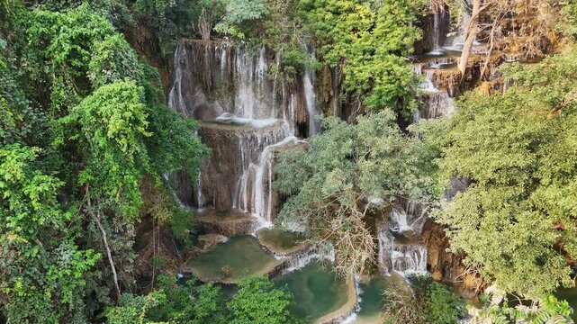 Aerial drone view of Kuang Si Waterfall main cascade near Luang Prabang, where multiple streams of water fall down limestone rock formations into turquoise pools surrounded by dense jungle
