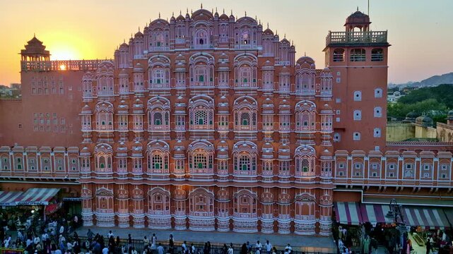 The sun sets behind the Hawa Mahal, casting a warm glow over its famous honeycomb facade. The grand pink sandstone structure towers above the lively evening crowds in Jaipur's historic center.