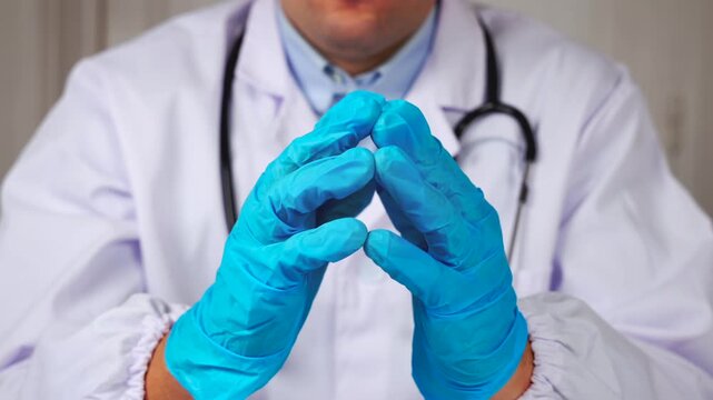 Un m&eacute;decin en blouse blanche et st&eacute;thoscope, avec des gants en nitrile bleu se tenant les mains.