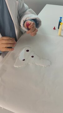 A young student wearing a white laboratory coat carefully drips red liquid colorant onto a smooth white slime base during a science experiment.