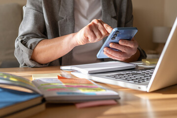 Businesswoman's hands holding and navigating a smartphone with a laptop, spiral notebook, and an open planner on a wooden desk, symbolizing remote work, financial management, and business planning