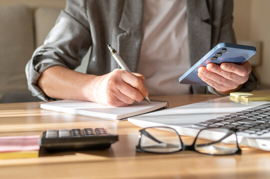 Business professional multitasking. Writing notes in a spiral notebook while holding a smartphone. With a calculator. Laptop. And eyeglasses on a wooden desk. Managing financial planning and budgeting