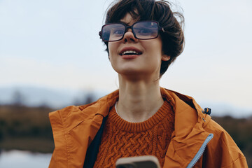 Young woman lifestyle outdoors wearing glasses and orange jacket smiles happily while holding a...